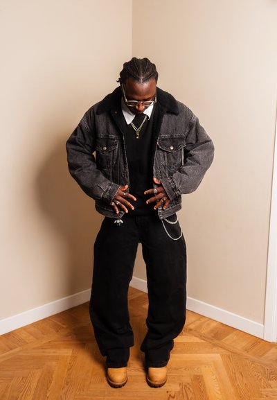 Hombre con cabello trenzado que lleva gafas, una chaqueta de mezclilla negra, pantalones negros, botas color canela y varios anillos, de pie sobre un suelo de madera en interiores.