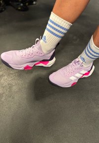 Persona que lleva zapatillas Adidas lavanda con rayas blancas y acentos rosas, combinadas con calcetines blancos que tienen rayas y logotipos azules.