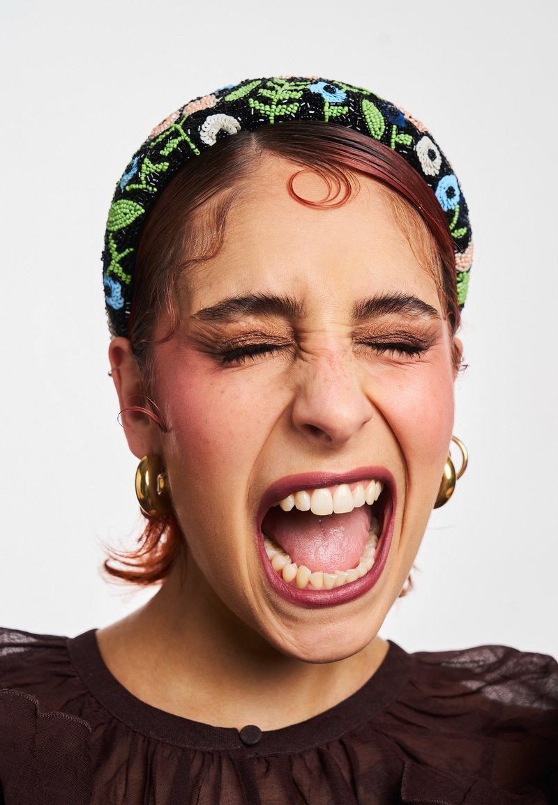 Fascia nera di perline con motivi floreali, indossata da una persona con capelli corti e rossastri, che sorride ampiamente mostrando i denti e indossando orecchini.