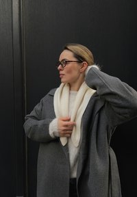 Grå ullkåpe med store slag over en hvit, fluffy genser; briller med skilpaddemønster; glatt hår; foran en mørk bakgrunn.