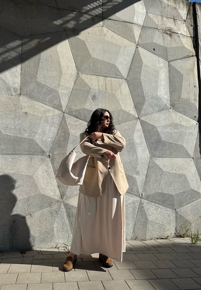 Blazer beige sobre un vestido blanco, sosteniendo un bolso gris claro. El fondo presenta una pared geométrica gris texturizada. La luz cálida del sol proyecta sombras.