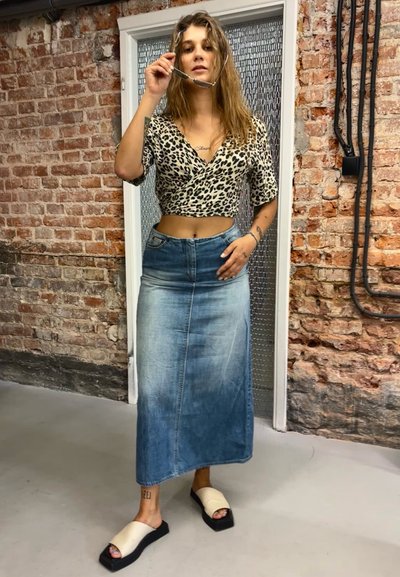 Top corto de estampado de leopardo, falda larga de mezclilla azul y sandalias de tiras color crema. Fondo de pared de ladrillo con suelos y accesorios industriales.