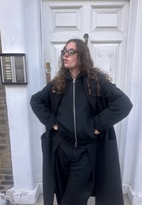 Manteau extérieur noir avec un sweat à capuche en dessous, debout devant une porte blanche. Texture filiforme notable et lunettes.
