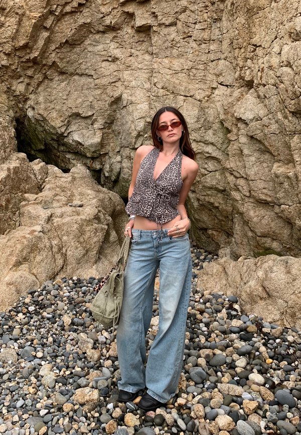 Top sin mangas de estampado de leopardo, jeans de pierna ancha azul claro y botas negras. Los accesorios incluyen un bolso verde y gafas de sol. Fondo de playa rocosa.