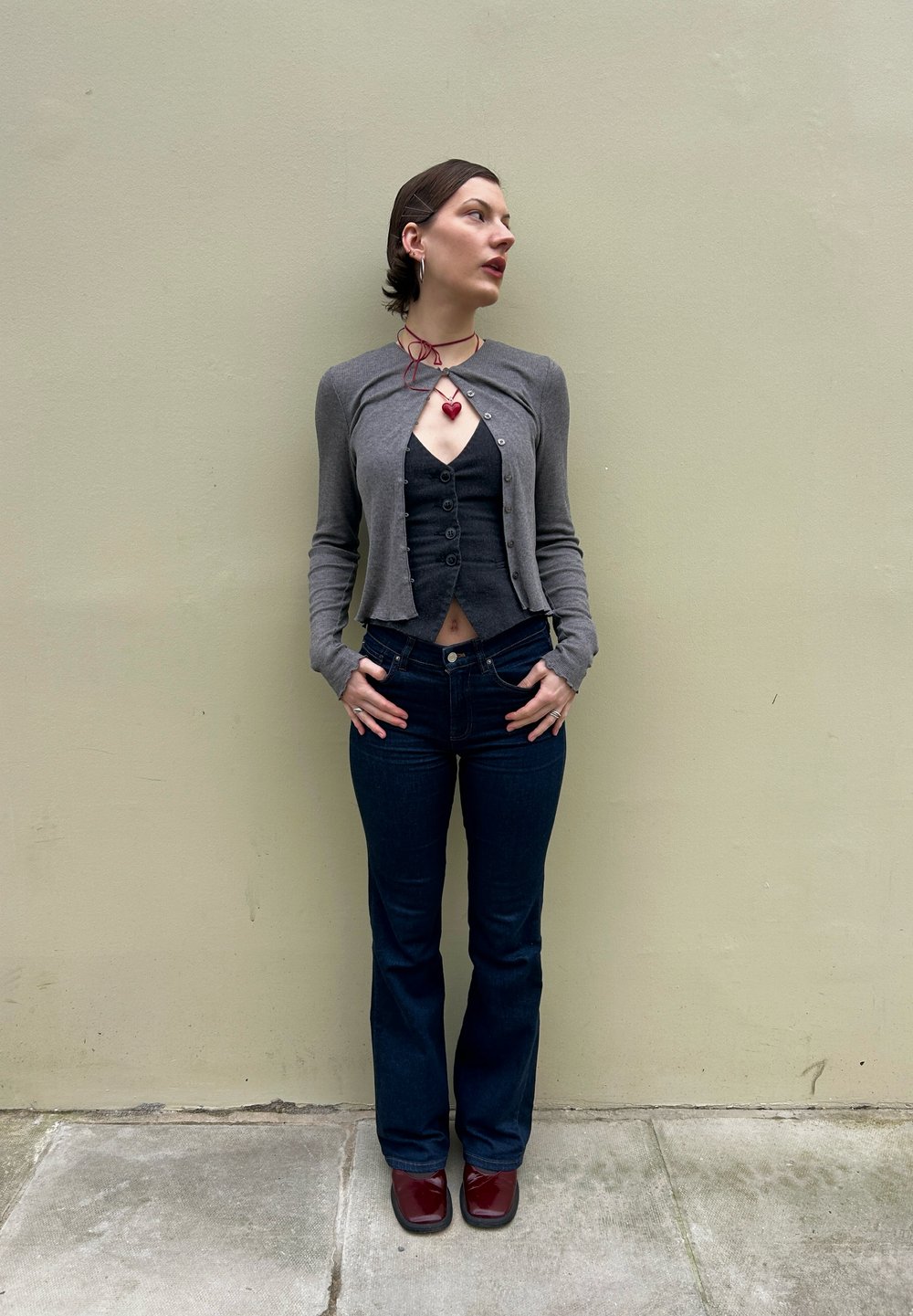 Cardigã cinza curto sobre uma blusa preta com botões, combinado com jeans escuros de boca de sino e sapatos burgundy; joias em camadas com um pendente em forma de coração.