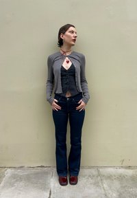 Cardigã cinza curto sobre uma blusa preta com botões, combinado com jeans escuros de boca de sino e sapatos burgundy; joias em camadas com um pendente em forma de coração.