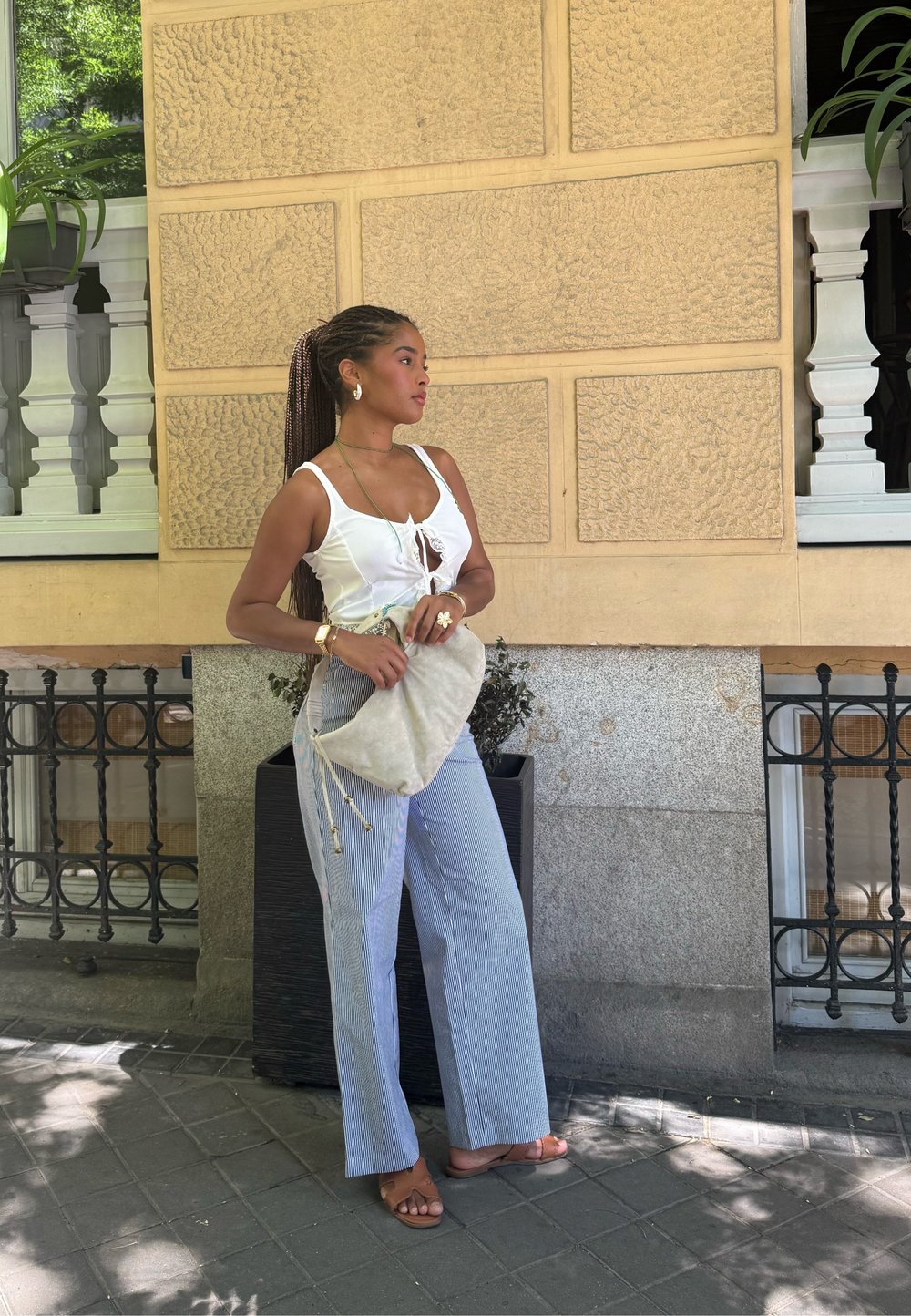 White tank top with a front tie, pinstriped wide-leg trousers, beige fabric bag with drawstrings, and brown sandals against a street backdrop.