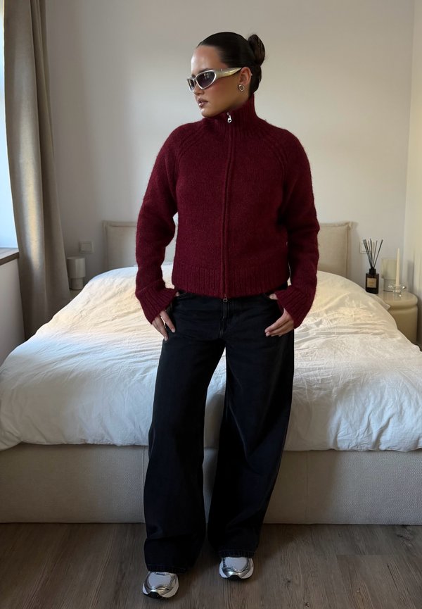 Una mujer luce en un dormitorio una sudadera burdeos con cremallera, pantalones negros anchos, gafas de sol y zapatillas deportivas.