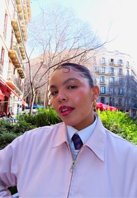 Jeune femme aux cheveux tressés et boucles d'oreilles en or, vêtue d'une veste rose et d'une cravate, en plein air dans un cadre urbain baigné de soleil avec des bâtiments et de la verdure.