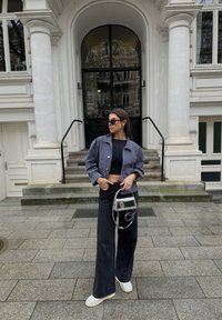 Pruhovaná džínsová bunda, čierny skrátený top, široké čierne nohavice, biele tenisky a priehľadná kabelka, umiestnené pred vstupom do klasickej budovy.