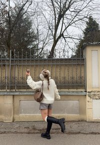 Kreemjakarva kudum tekstuursete detailidega, hallid lühikesed püksid, põlveni ulatuvad mustad saapad ja pruun käekott. Väljas taustal puud.