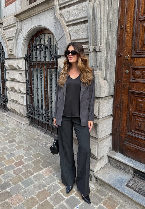 Femme portant des lunettes de soleil noires, un blazer gris, un haut noir et un pantalon à jambes larges, debout sur un pavement en pierre près de fenêtres en fer ornées et d'une porte en bois.