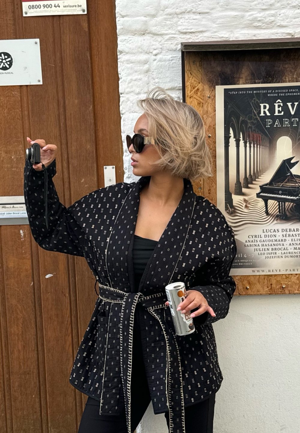 Svart mønstrete jakke med en omslagsdesign, teksturert stoff og kontrastfarget hvit søm. Båret over en svart topp, holder en drink og et kamera.