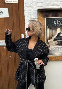 Svart mønstrete jakke med en omslagsdesign, teksturert stoff og kontrastfarget hvit søm. Båret over en svart topp, holder en drink og et kamera.