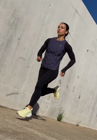 Odzież sportowa z ciemnofioletową kamizelką na czarnych długich rękawach, w połączeniu z czarnymi legginsami i jasnobłękitnymi butami do biegania.