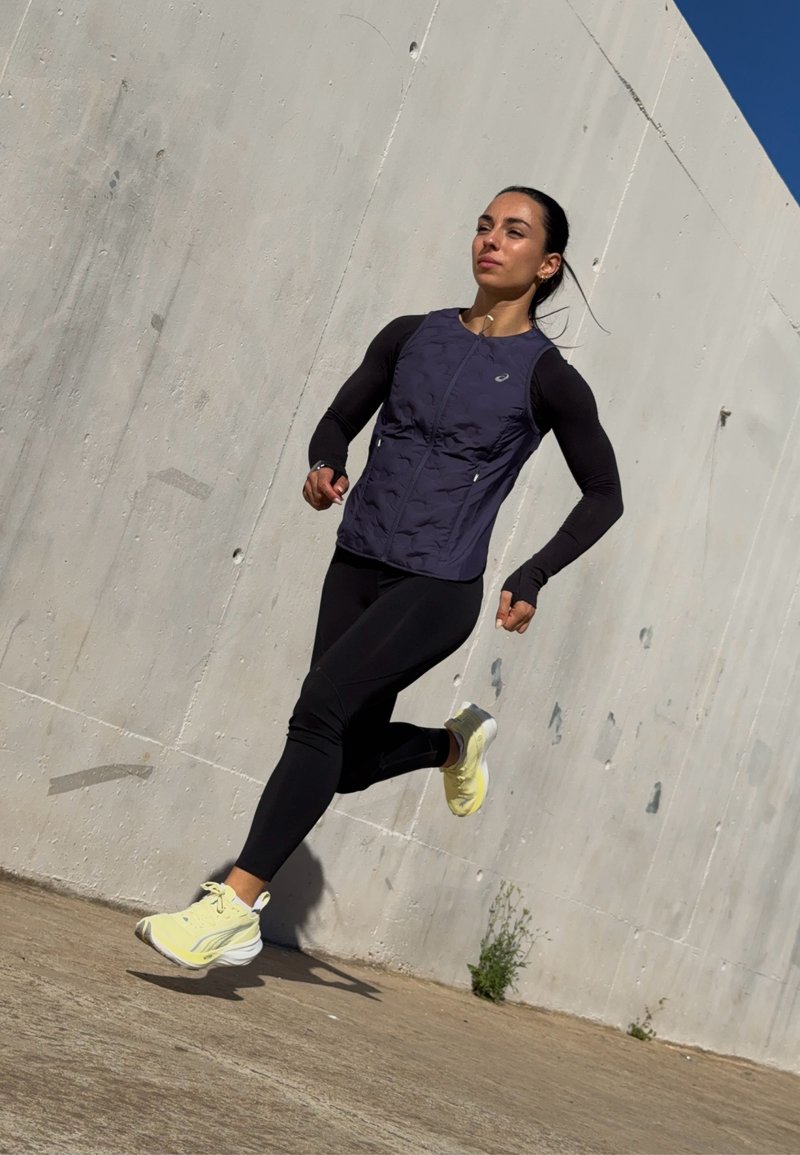 Athletic apparel featuring a dark purple vest over black long sleeves, paired with black leggings and light yellow running shoes.