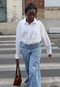 Biała koszula na guziki, jasnoniebieskie szerokie dżinsy, trzymająca brązową torbę typu tote, przechodząca przez pasy na przejściu dla pieszych, w tle teksturowana ściana.