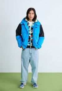 Blå och svart jacka med huva, lager på lager över en flerfärgad mönstrad fleece, tillsammans med ljusblå jeans och sportiga skor.