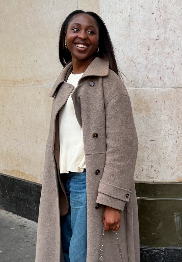 Donna sorridente che indossa un lungo cappotto beige, un maglione bianco e jeans blu, in piedi contro un muro di pietra chiara, con una borsa a catena in mano.