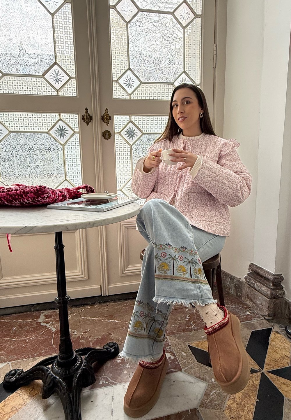 Lys rosa quiltet jakke med flæse detaljer, blå denim jeans med blomstermønster, brune slip-on sko, hvid kop i hånden, marmor bord.