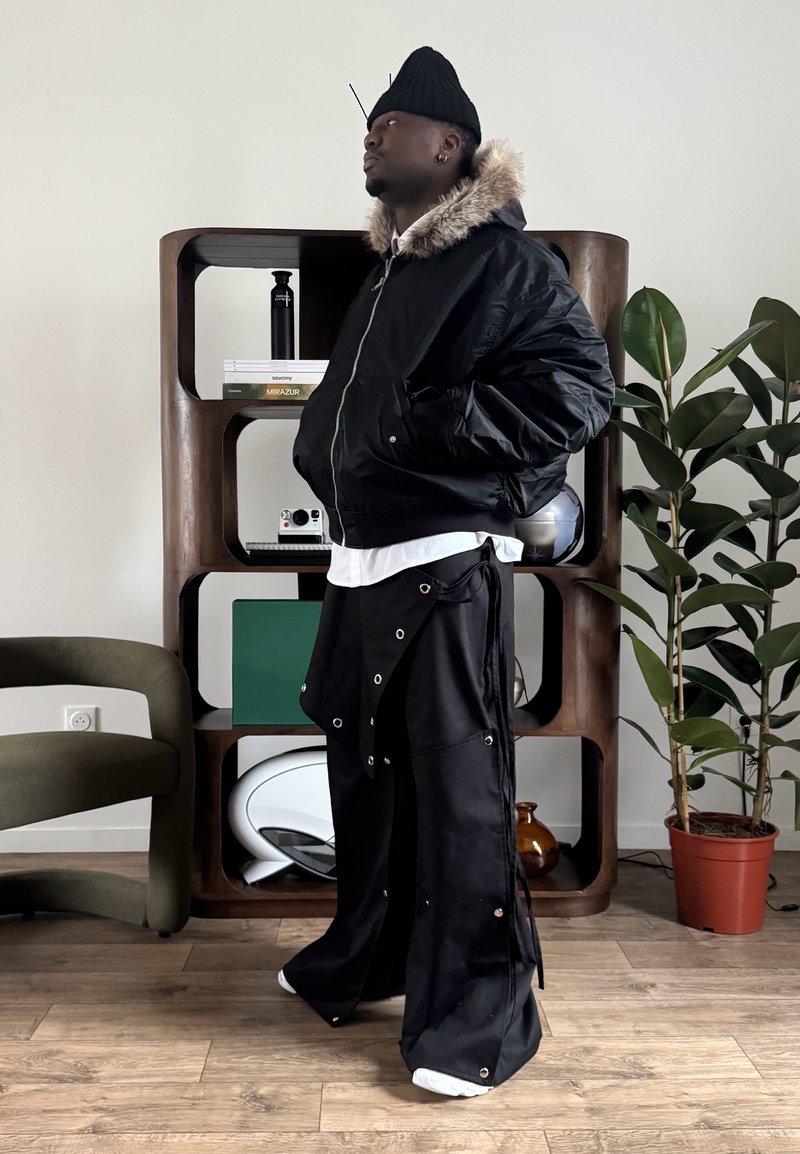 Veste bomber noire avec capuche doublée de fourrure, associée à un pantalon noir avec détails en œillets métalliques. Le mannequin se tient à côté d'une décoration moderne.