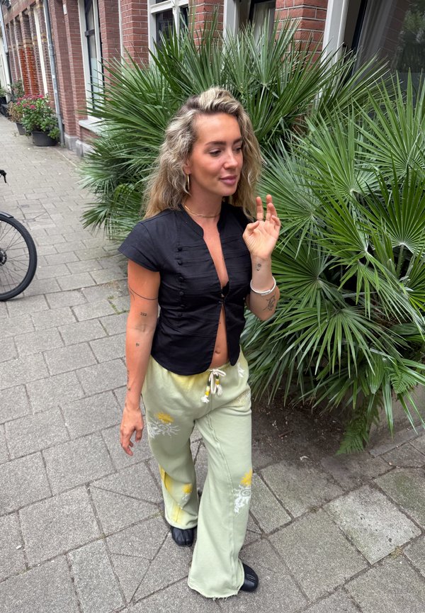 Femme aux cheveux blonds marche sur un trottoir en ville à côté de plantes de palmier vertes, portant un haut noir et un pantalon vert clair à motifs floraux.