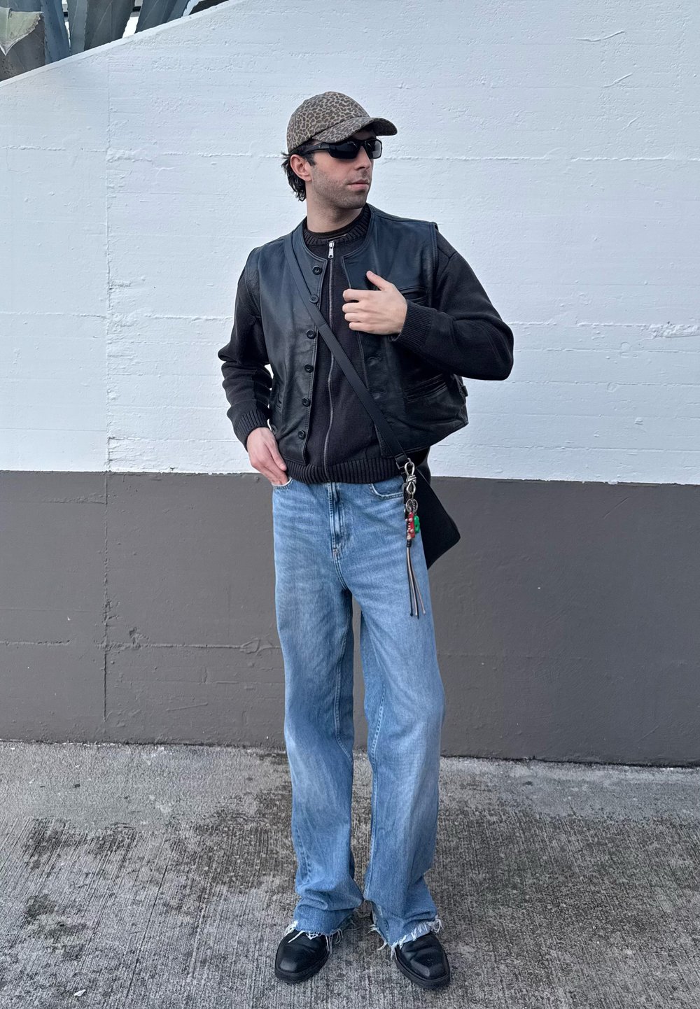 Μαύρο δερμάτινο γιλέκο πάνω από ένα σκούρο πουλόβερ, ανοιχτό μπλε παντελόνι σε φαρδιά γραμμή και μαύρα παπούτσια με τριμμένα άκρα. Τα αξεσουάρ περιλαμβάνουν μια καπέλο με σχέδιο και μια τσάντα.