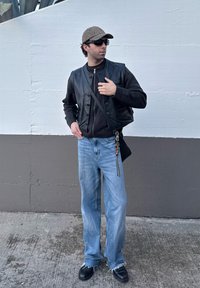 Μαύρο δερμάτινο γιλέκο πάνω από ένα σκούρο πουλόβερ, ανοιχτό μπλε παντελόνι σε φαρδιά γραμμή και μαύρα παπούτσια με τριμμένα άκρα. Τα αξεσουάρ περιλαμβάνουν μια καπέλο με σχέδιο και μια τσάντα.