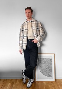 Άνδρας με μπεζ καρό σακάκι, μπεζ πουλόβερ, μαύρο παντελόνι και λευκά αθλητικά παπούτσια, στέκεται με το ένα πόδι σταυρωμένο δίπλα σε κορνιζαρισμένη αφηρημένη γραμμική τέχνη.