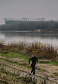 Persoon in zwarte sportkleding die jogt op een onverharde weg langs een rivier met bladloze bomen en een groot stadion op de achtergrond op een bewolkte dag.