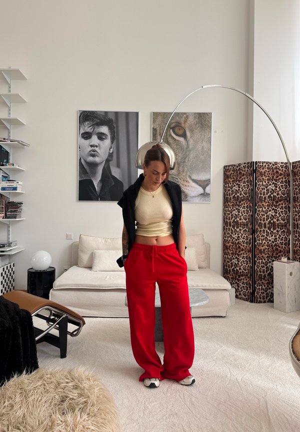 Vrouw in rode broek en beige cropped top staand in een stijlvolle woonkamer met luifel in luipaardprint, moderne meubels en zwart-wit wandfoto's.