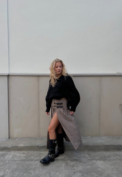 Suéter negro de punto con mangas abullonadas, falda maxi de cuadros con dos hebillas y botas de cuero negras hasta la rodilla con acentos metálicos.