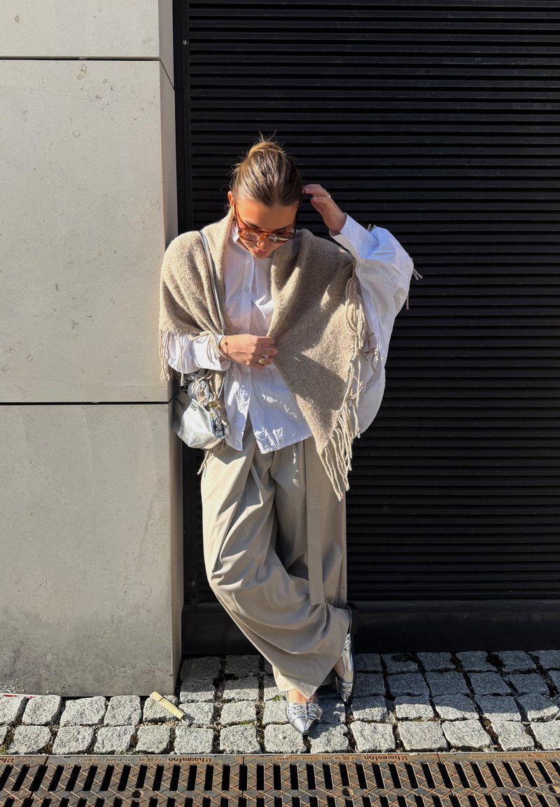 Femme portant des lunettes de soleil, une chemise blanche, un châle beige, un pantalon ample et des chaussures argentées, appuyée contre un mur dans une rue pavée sous le soleil.