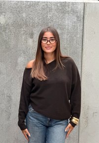 Jonge vrouw met lang steil haar en een bril, gekleed in een zwart trui met schouders bloot, blauwe spijkerbroek en grote gouden armbanden tegen een betonwand.