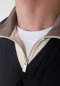 Veste noire zippée avec une doublure intérieure beige et un col texturé. T-shirt blanc visible en dessous avec un col rond.