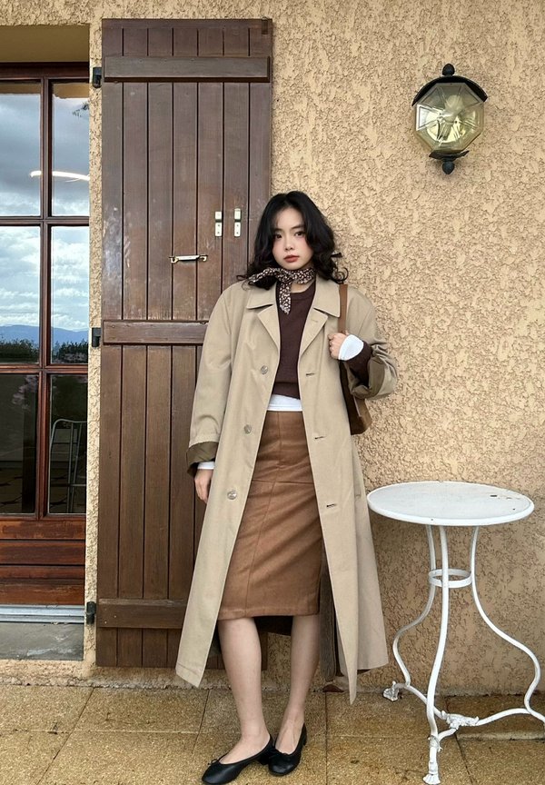 Une femme portant un trench beige et une jupe brune pose à côté d’une porte en bois, avec une petite table blanche contre le mur crépi.