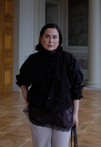 Veste coupe-vent noire à col montant, portée sur un haut noir orné de dentelle. Pantalon de couleur claire et sac à main noir visibles.