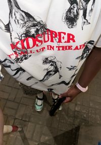 White T-shirt adorned with black graphic sketches of figures, complemented by red text stating "KIDSUPER" and "IT'S ALL UP IN THE AIR," featuring short sleeves.