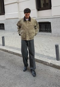 Veste en toile vert olive avec un col marron, des boutons sur le devant et deux poches latérales, assortie à un pantalon à plis foncé et des chaussures noires.