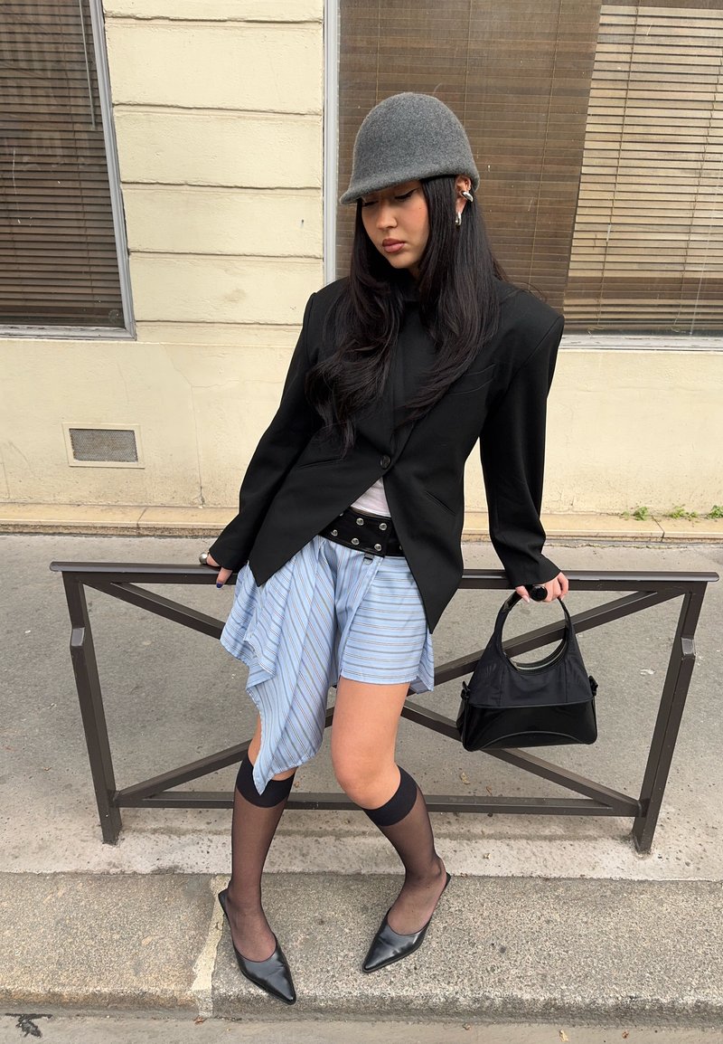 Jeune femme portant un chapeau gris, un blazer noir, une jupe asymétrique rayée, des chaussettes noires transparentes montant jusqu'aux genoux et des talons noirs, tenant un sac à main noir.