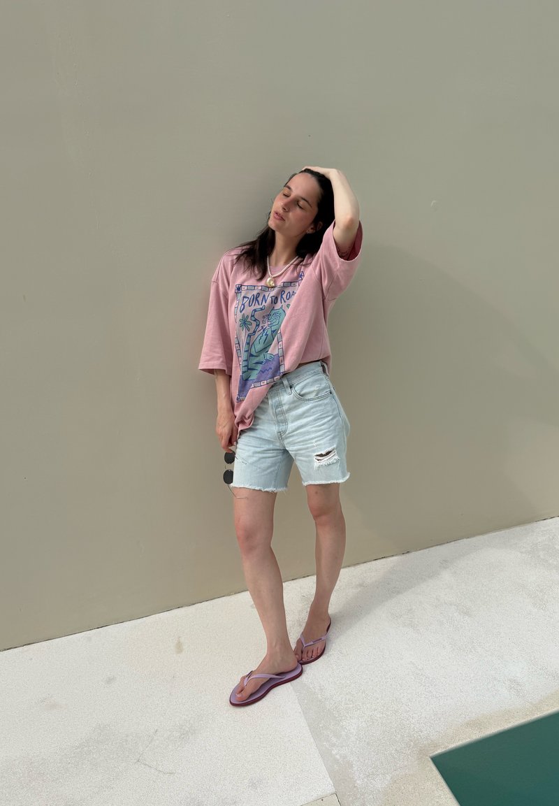 Kvinde i pink grafisk t-shirt, lyse slidte jeansshorts og lilla klipklapper står barfodet på hvidt gulv ved beige væg og holder solbriller.