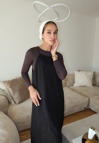 Μαύρο ριγέ φόρεμα τοποθετημένο πάνω από ένα εφαρμοστό καφέ τοπ, με άσπρη κορδέλα και κοσμήματα σε χρυσό τόνο, σε φόντο από μια φωτεινή σάλα καθιστικού.