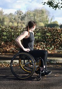 Fauteuil roulant manuel avec cadre noir et accents jaunes. Utilisateur portant un haut sans manches noir et un pantalon, positionné sur un chemin entouré de feuillage.