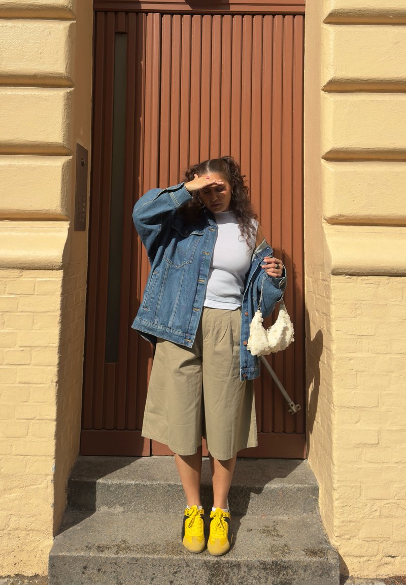 Femme se protégeant les yeux du soleil, debout sur des marches devant une porte marron, portant une veste en denim, un short beige, un haut blanc et des chaussures jaunes.