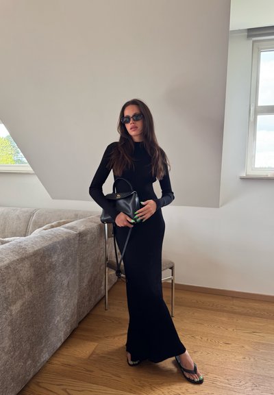 Vestido negro de manga larga con un corte ajustado. Lleva un bolso negro. Usa gafas de sol negras y chanclas con acentos verdes.