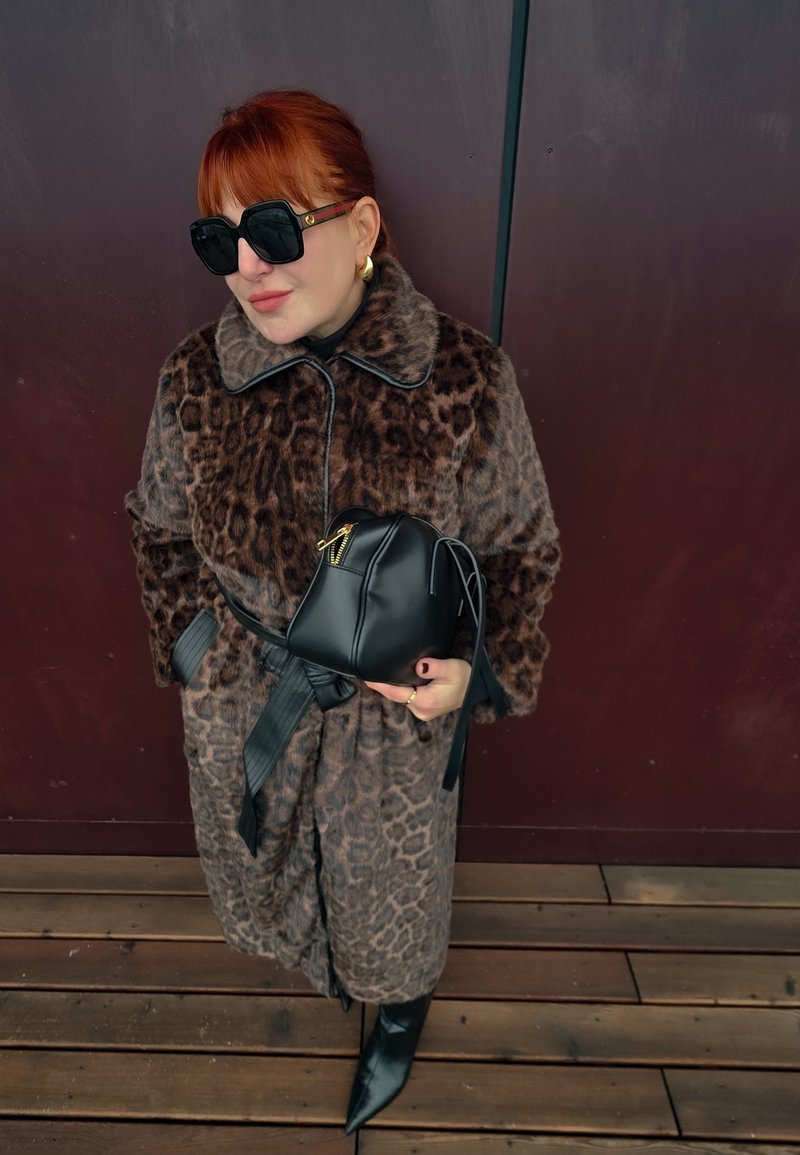 Manteau en fourrure synthétique à imprimé léopard avec accents noirs, ceinturé, associé à un sac en cuir noir et des bottes pointues noires, porté avec des lunettes de soleil oversize.