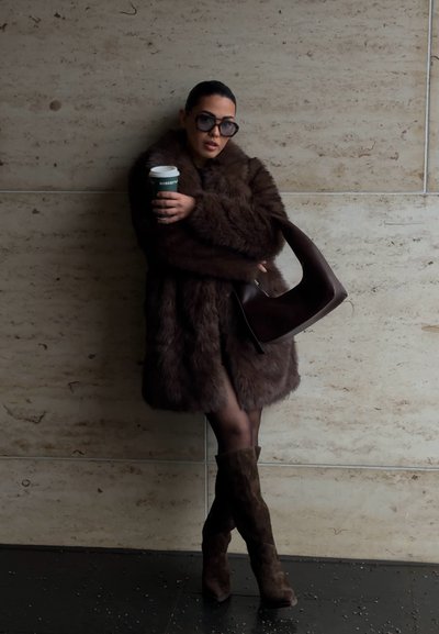 Abrigo de piel sintética marrón, bolso de mano marrón oscuro, medias oscuras, botas de ante marrón hasta la rodilla. Sosteniendo una taza de café, de pie contra una pared clara.
