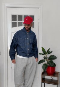 Chaqueta de denim azul con dos bolsillos en el pecho, pantalones deportivos grises y una gorra roja con un logo de corazón, frente a una pared blanca y una planta verde.