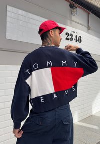Σκούρο μπλε πλεκτό πουλόβερ με μεγάλη κόκκινη και λευκή γραφική εκτύπωση που αναγράφει "TOMMY JEANS" με λευκά γράμματα στην πλάτη; χαλαρή εφαρμογή.
