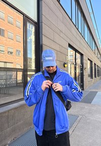 Μπλε φανέλα με φερμουάρ και λευκές λωρίδες, πάνω από μαύρη μπλούζα. Ο φορέας προσαρμόζει μία τσάντα χιαστί, φορώντας μπλε καπέλο με λογότυπο "Supreme".